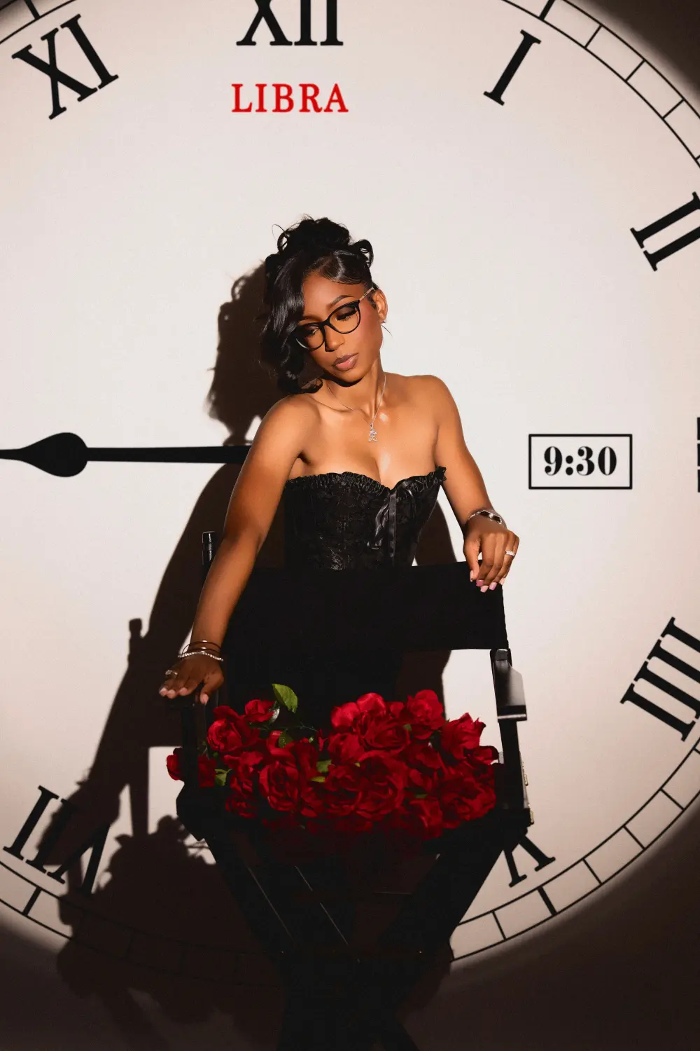 Woman in glasses and a black corset seated with red roses in front of a giant projected clock
