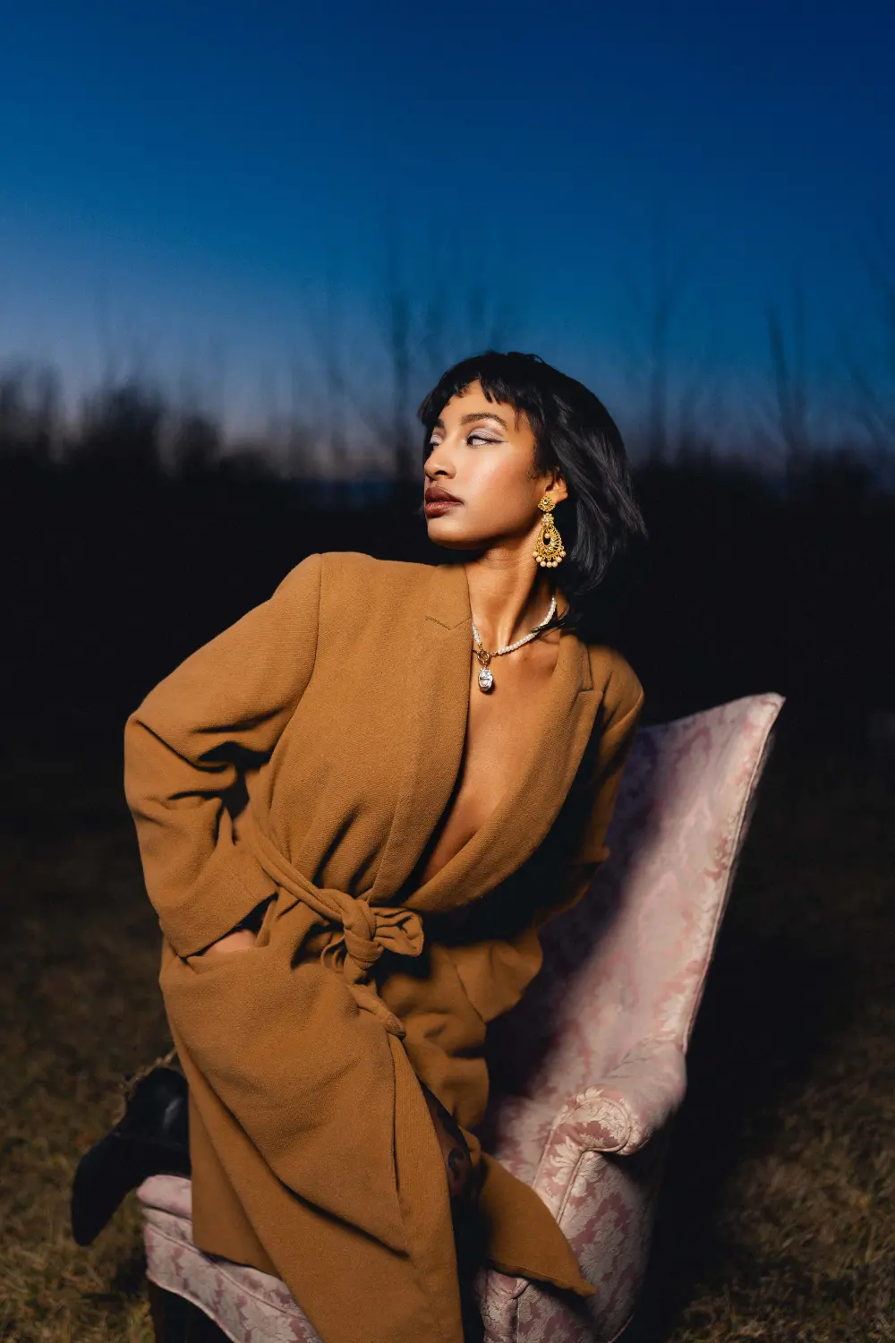 Woman in a camel coat on an antique armchair in a field at blue hour