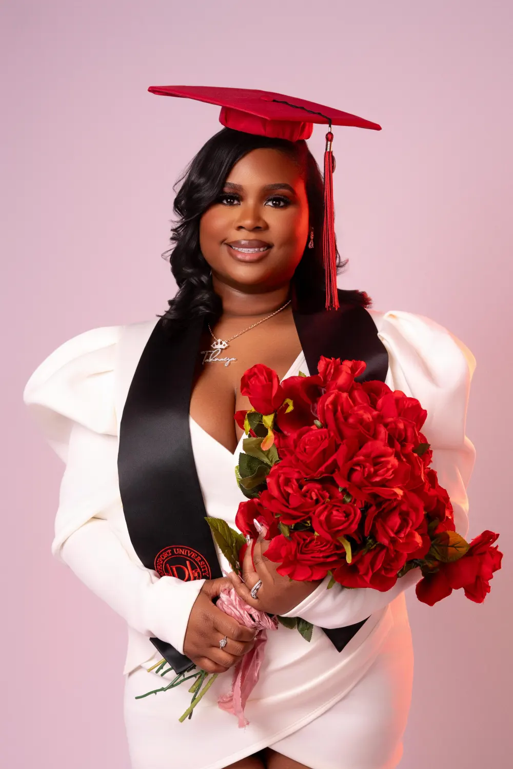 Graduate in a red cap, white blazer and black stole holding red roses against a pink backdrop
