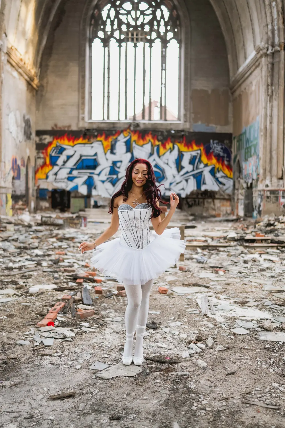 Woman in a white corset and tutu standing in the rubble of an abandoned church, graffiti and a tall arched window behind her