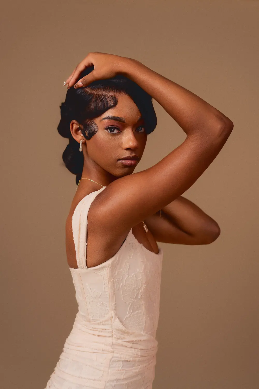 Woman in a cream dress with one arm over her head, looking over her shoulder against a tan backdrop