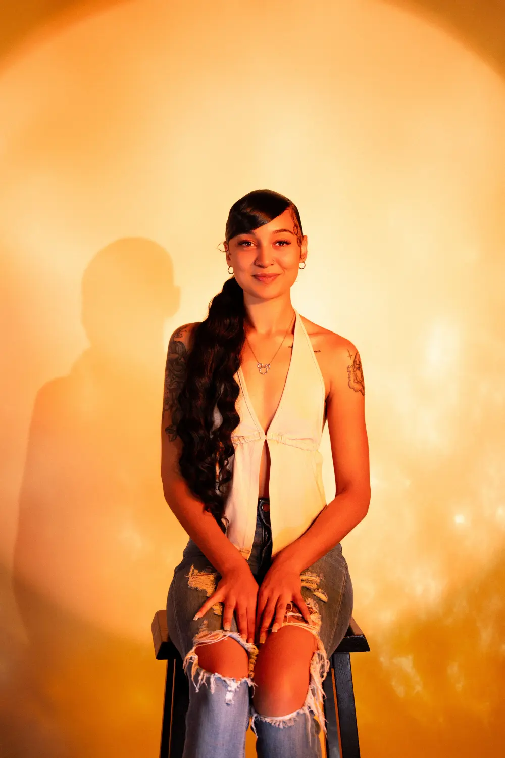Woman in a white halter top and ripped jeans seated on a stool against a glowing amber backdrop