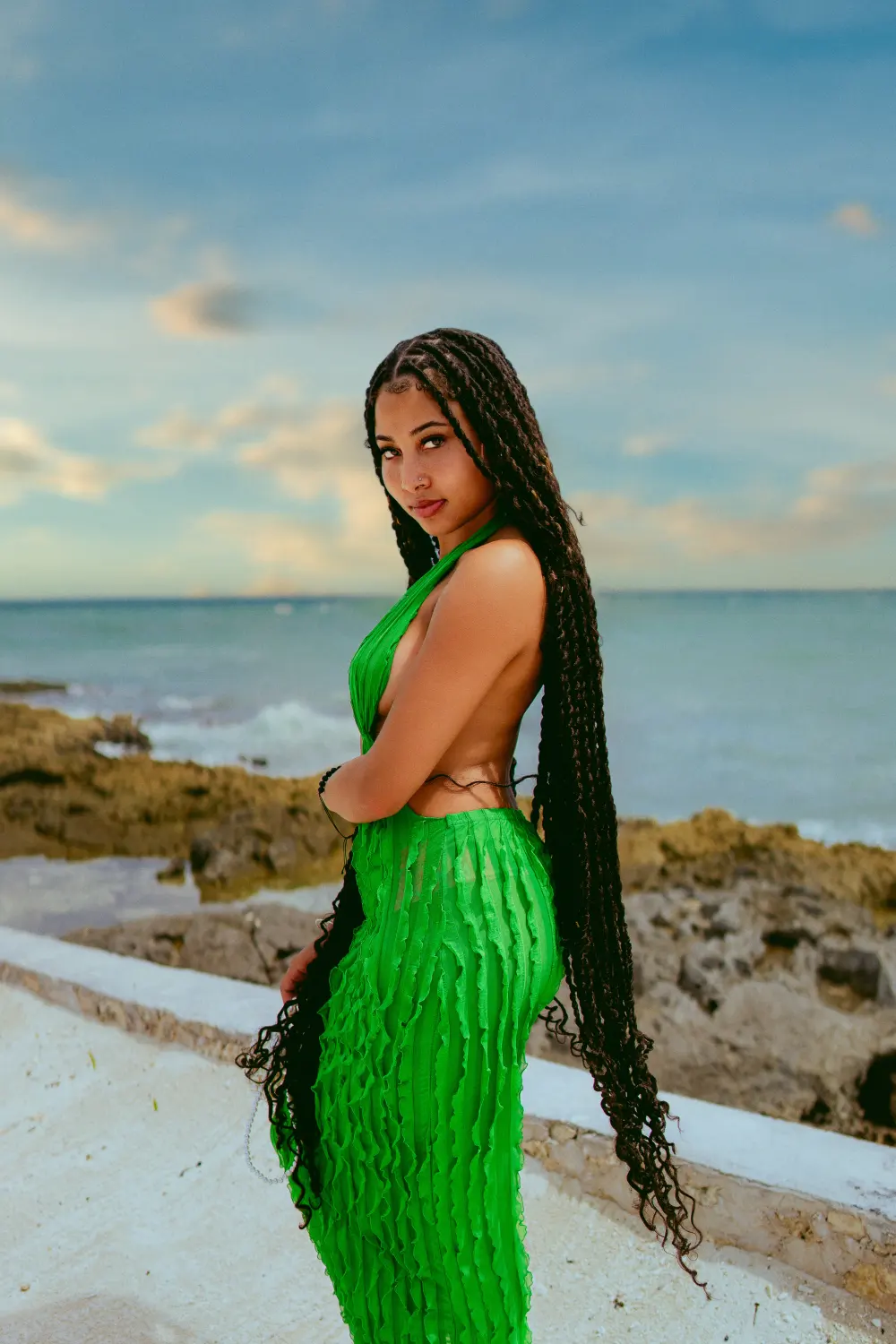 Woman in a green knit dress with long braids standing on beach rocks by the ocean