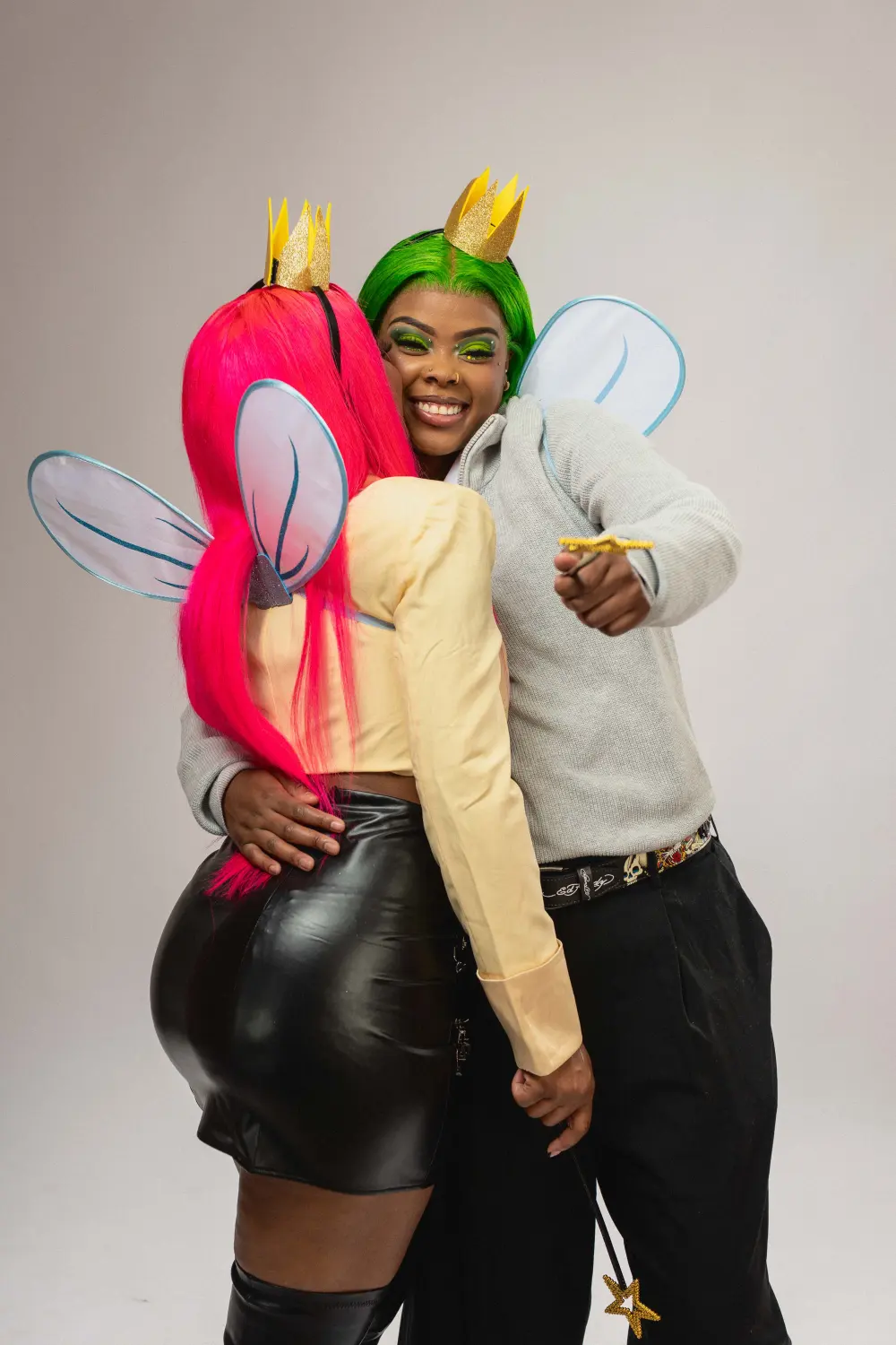 Couple dressed as cartoon fairy godparents with pink and green hair, crowns and wings, hugging in the studio