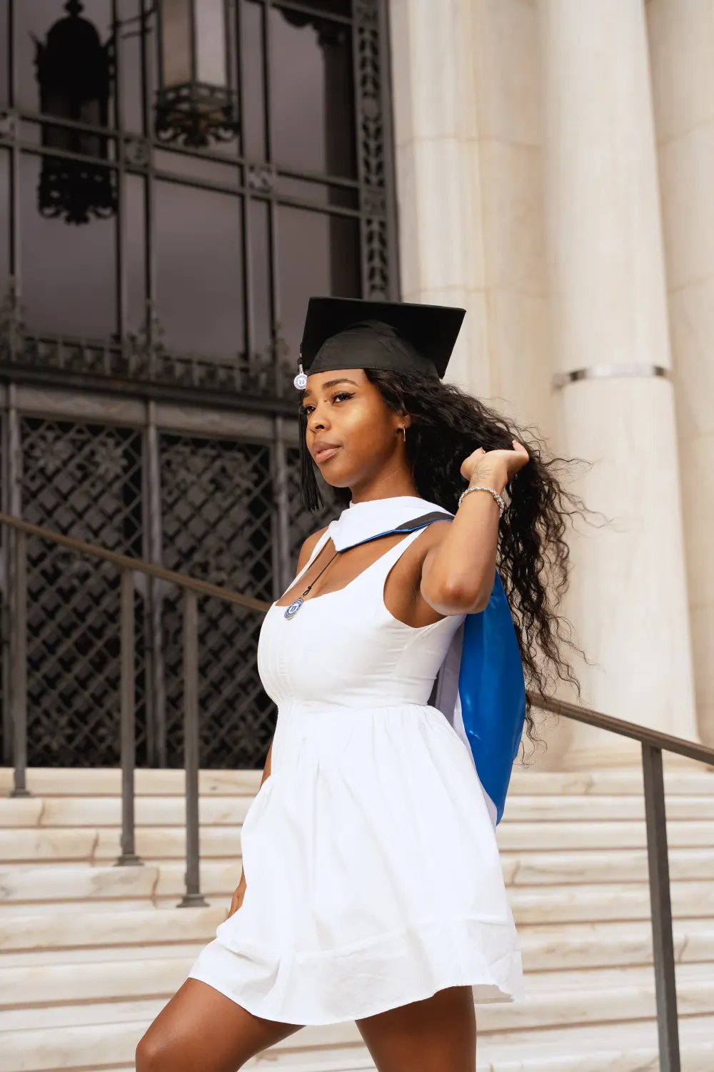 Graduate in a white dress and cap on marble steps in front of tall ornate doors, a blue and white stole over her shoulder