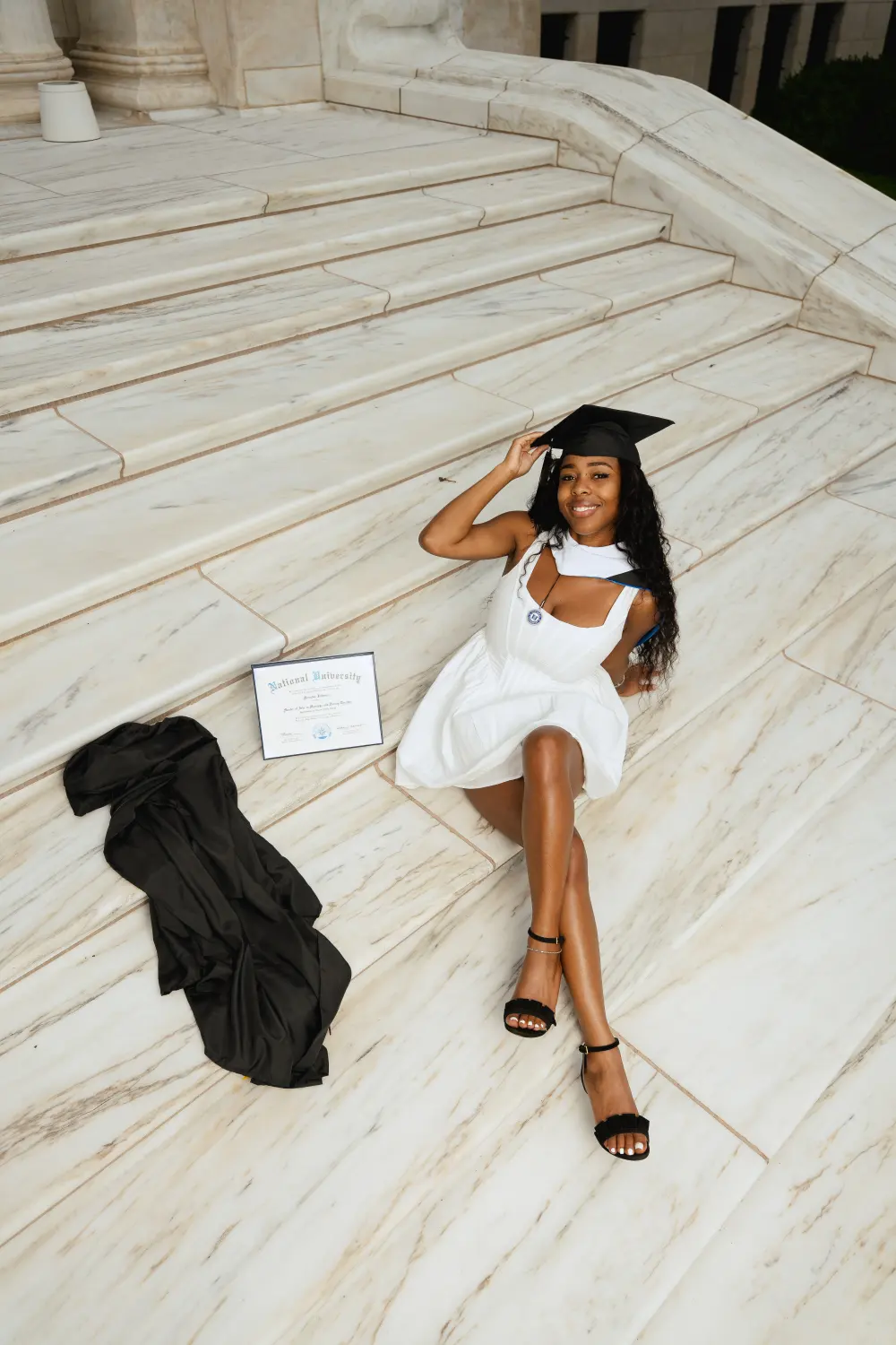 Graduate sitting on wide marble steps seen from above, her gown and diploma laid out beside her