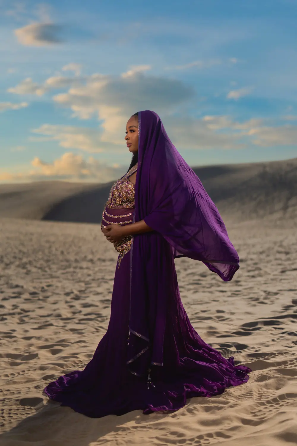 Pregnant woman in a flowing purple gown and veil standing on sand dunes under a blue sky