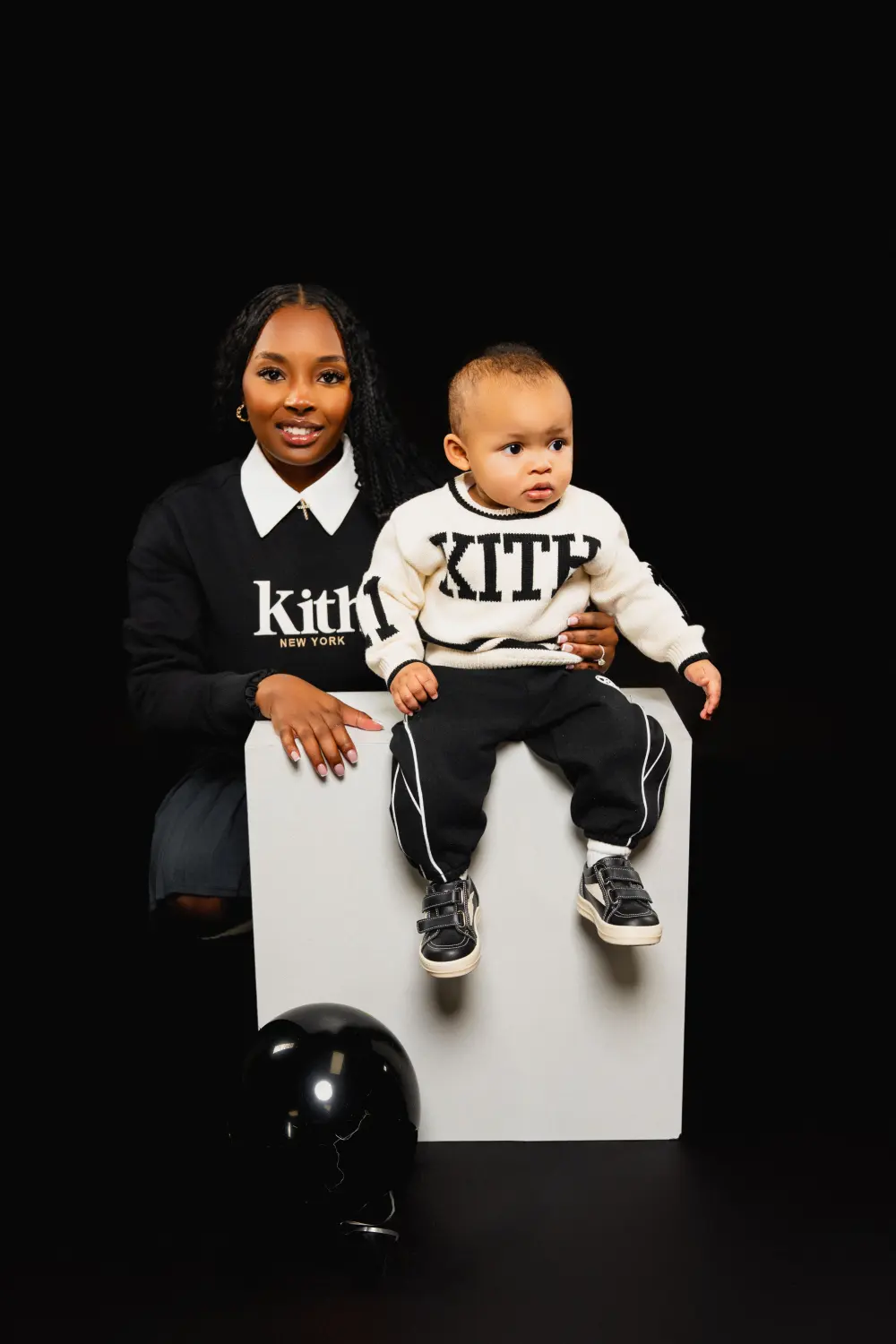 Mother and baby in matching black and white sweaters posing on a white cube against a black backdrop