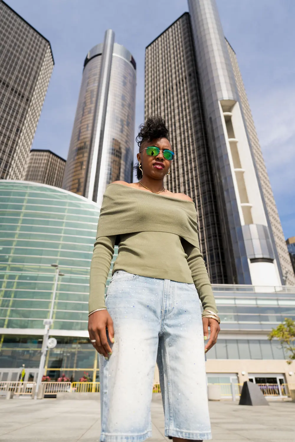 Woman in an olive off-shoulder top and light denim standing beneath the Renaissance Center towers